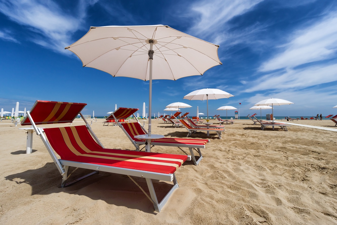 Sdraio rosse a righe e ombrelloni bianchi su una spiaggia sabbiosa sotto un cielo soleggiato.